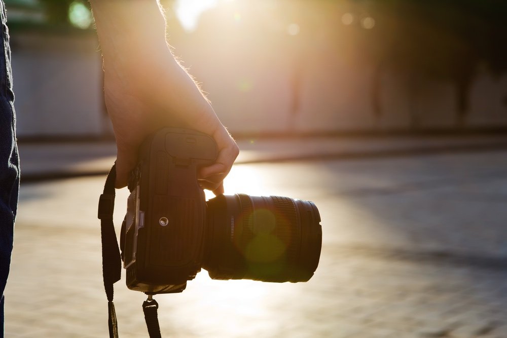 Photographer walking on pavement while holding his professional camera with lens attached. Strong sunlight in background.